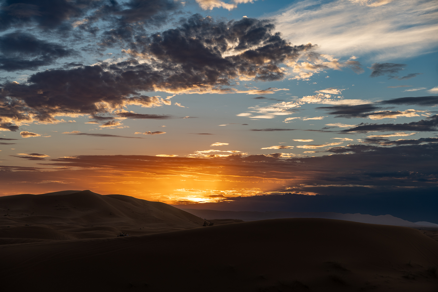 Dunas del desierto de Merzouga, Marruecos, recortadas en silueta bajo nubes oscuras y un cielo anaranjado en el horizonte.