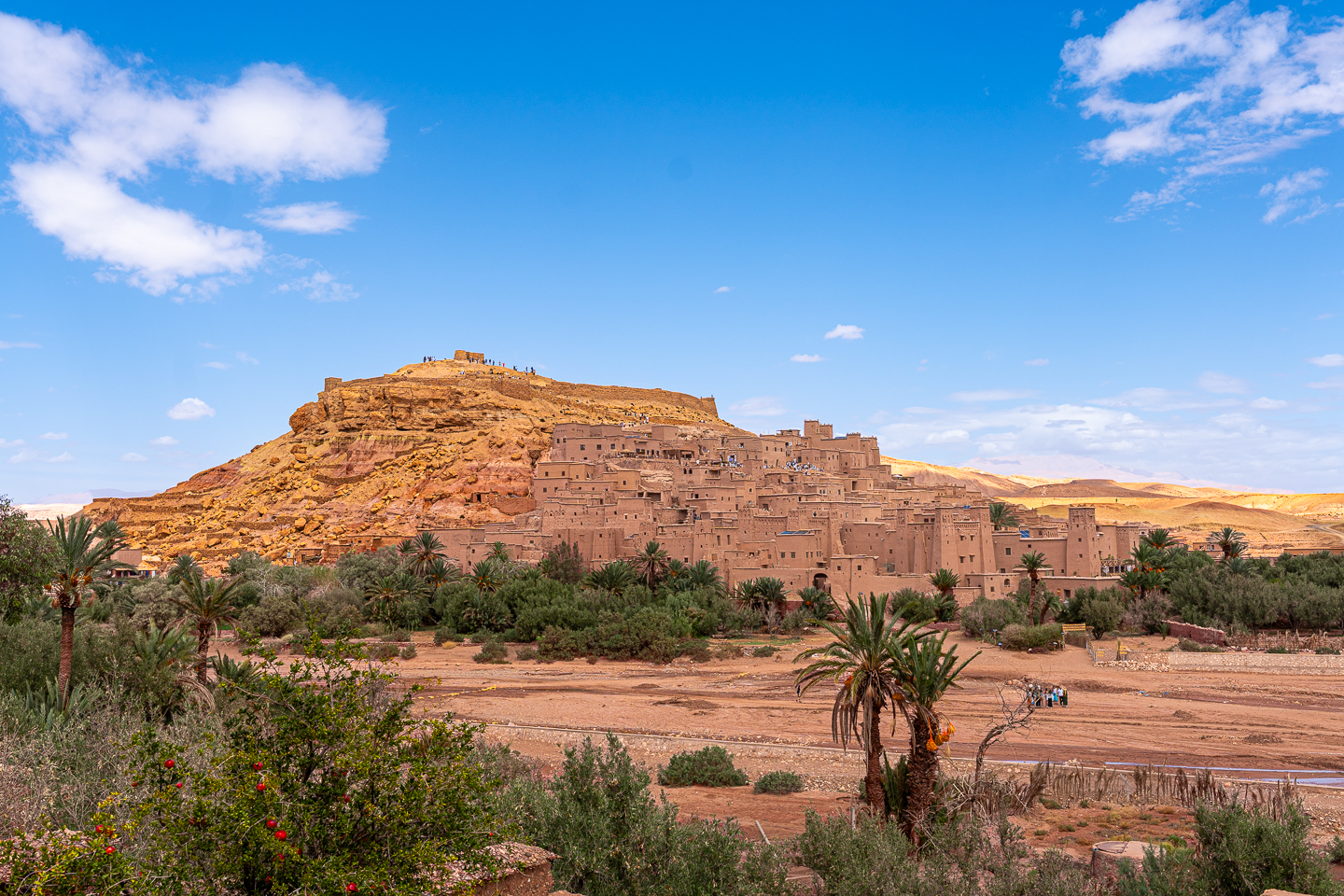 Earthen buildings and towers of Ait Ben Haddou, Morocco, rising up a rocky hillside above palm trees and a wide riverbed.