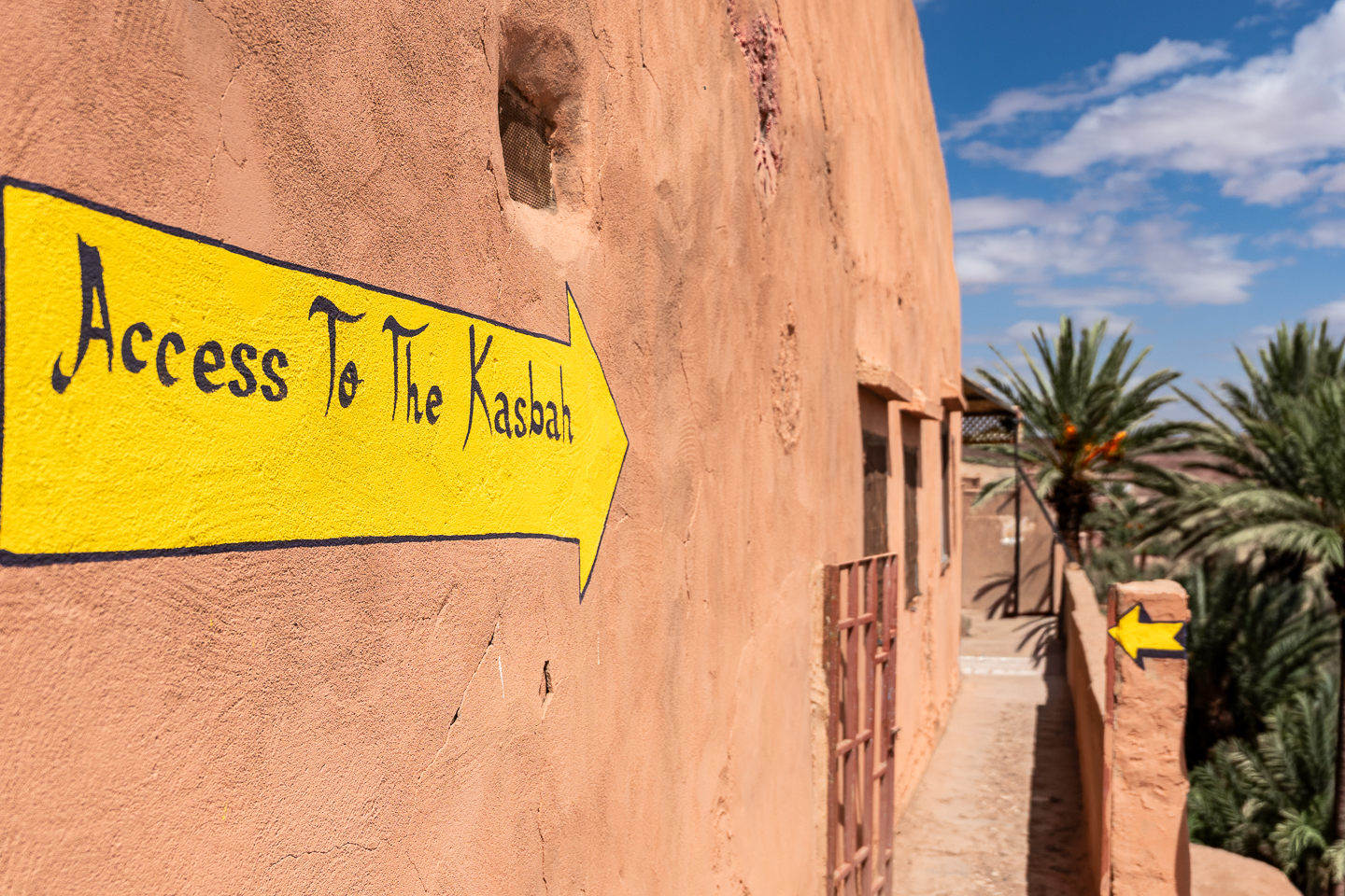 Yellow arrow reading “Access To The Kasbah” on an earthen wall beside a narrow walkway and palm trees.