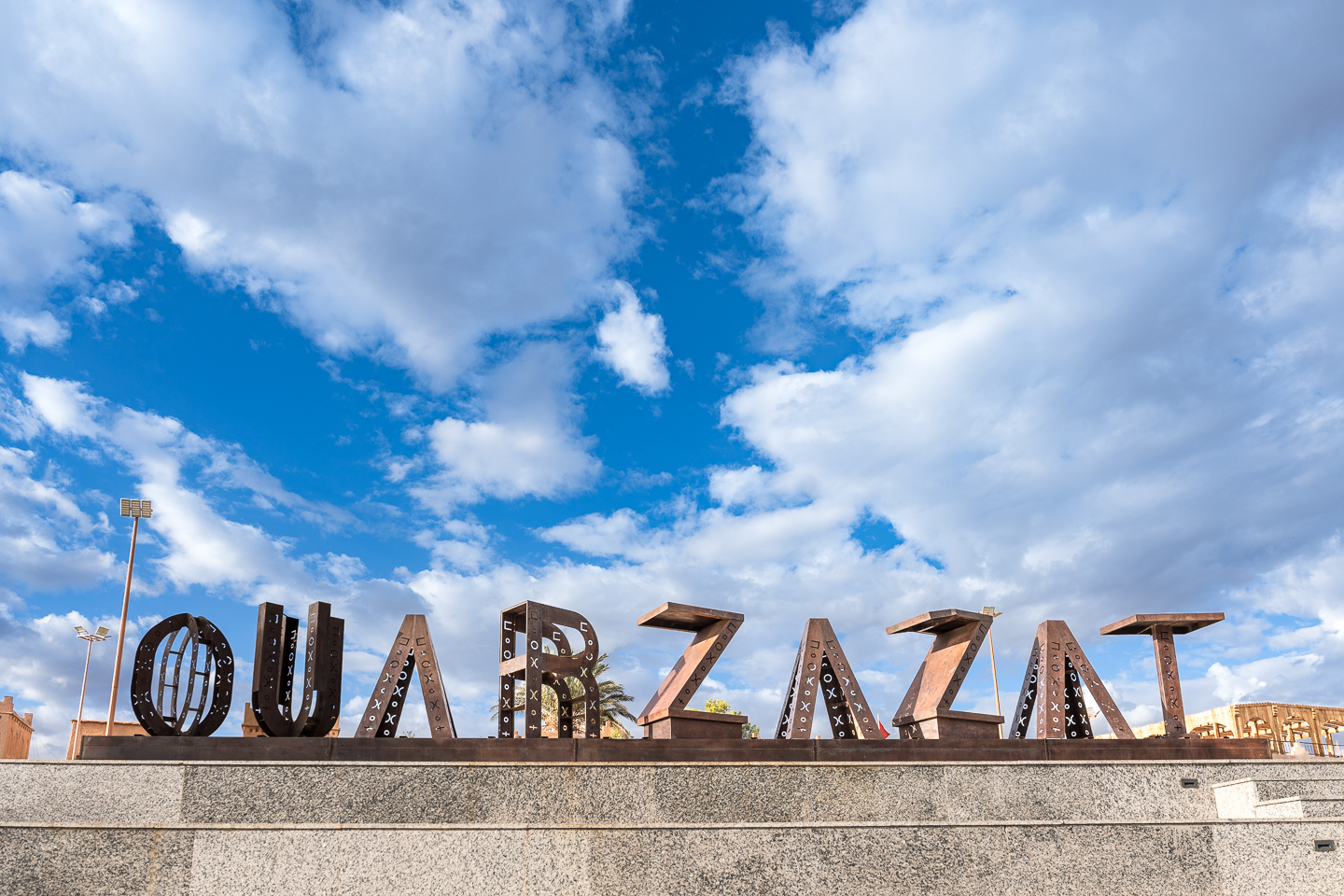 Large metal letters spelling “OUARZAZAT” on a raised stone platform beneath a blue sky filled with white clouds.