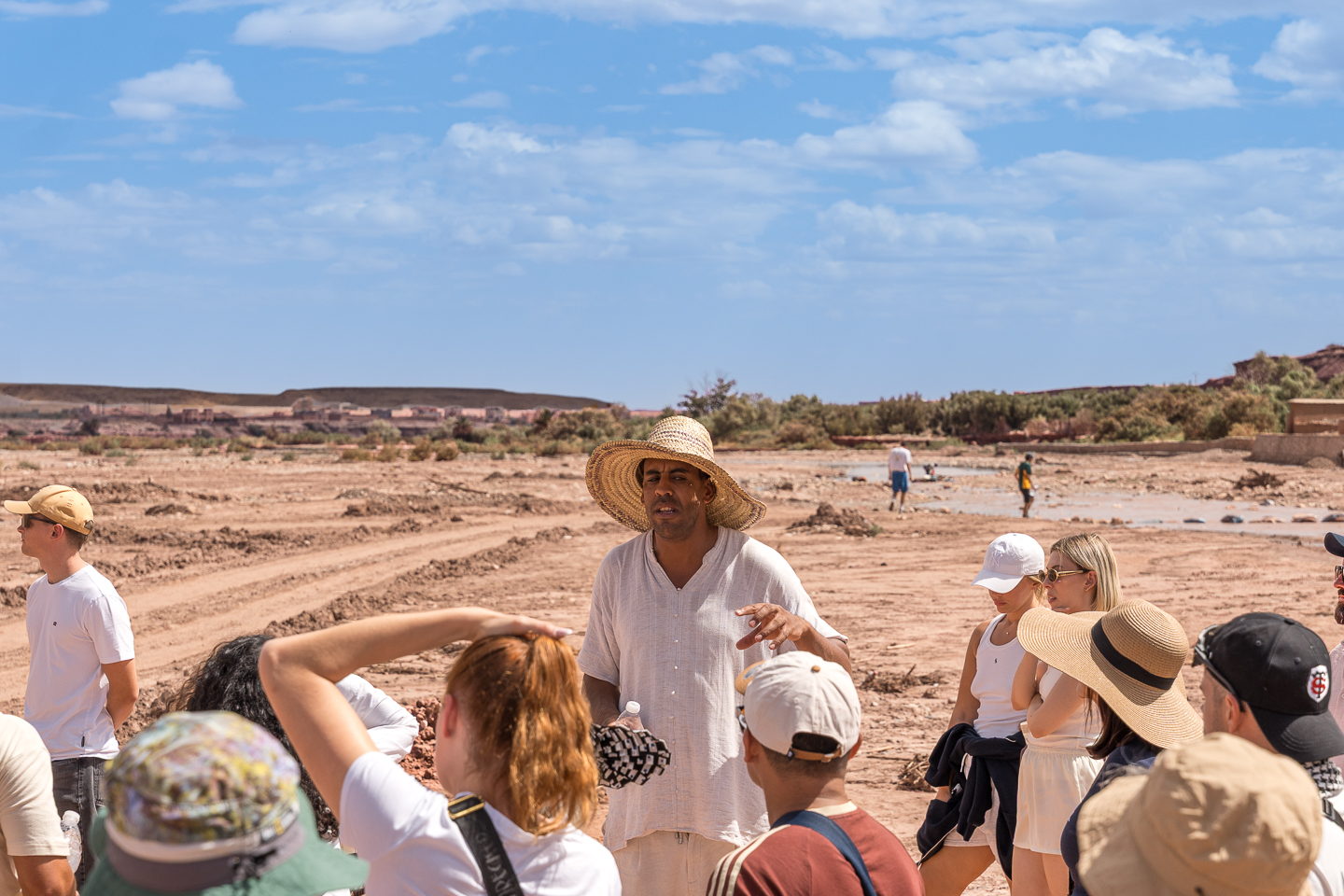 A guide wearing a straw hat speaking to a group of visitors beside a mostly dry riverbed.