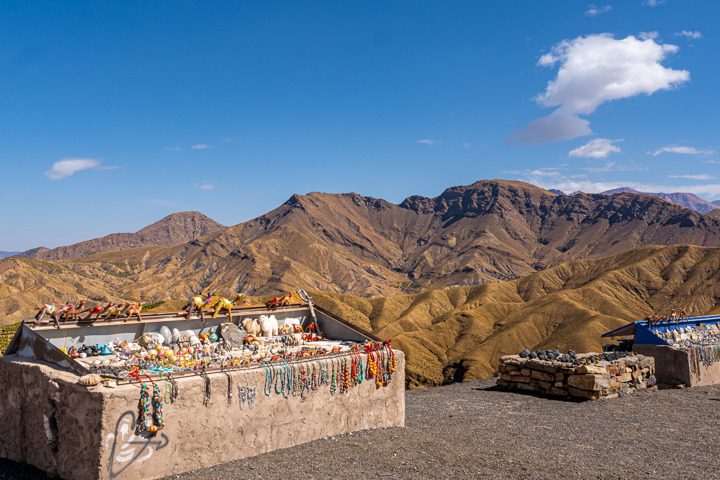 Souvenir stalls displaying colorful necklaces, stones, and camel figurines against rugged mountains at Tizi n’Tichka, Morocco.