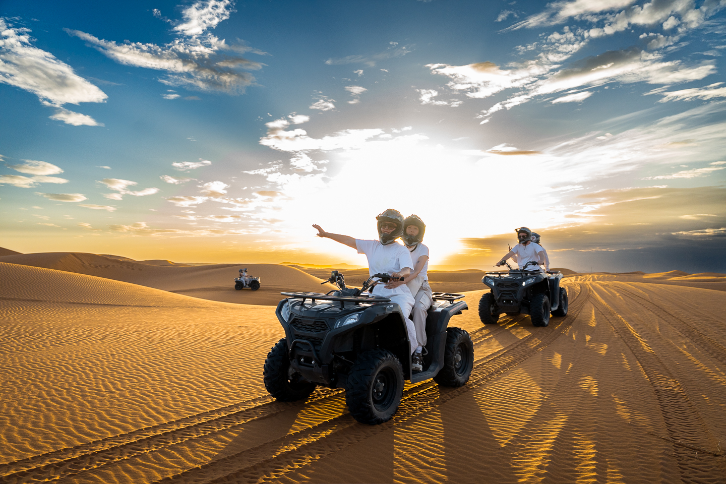 Helmeted riders on quad bikes crossing golden sand dunes in Merzouga, Morocco, with the low sun casting long shadows across the sand.