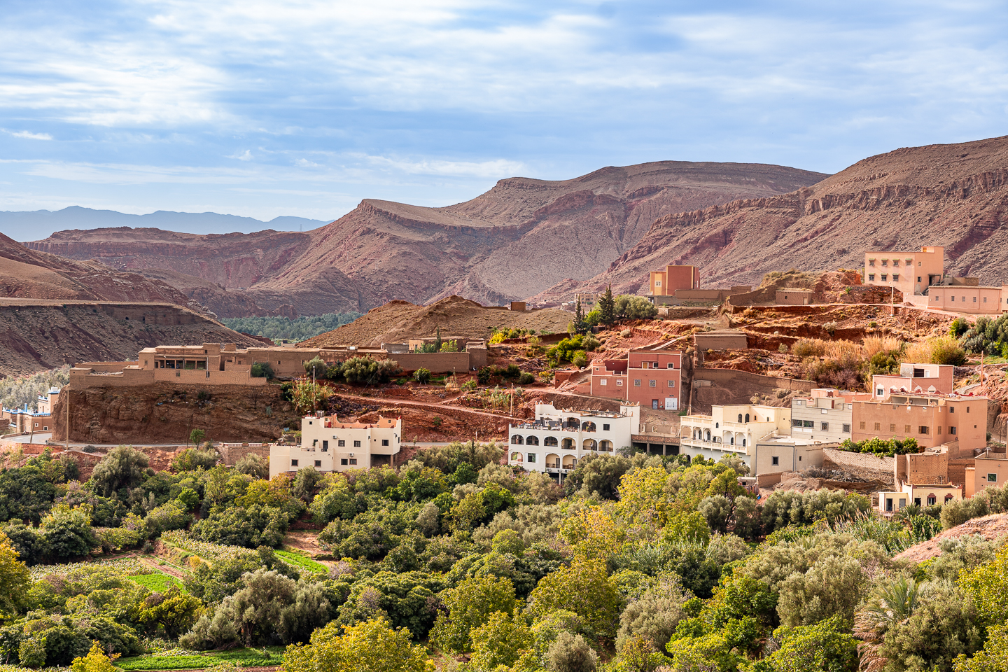 Pink and cream village buildings overlooking green gardens and trees, with rugged, dry mountains rising behind them in Morocco.