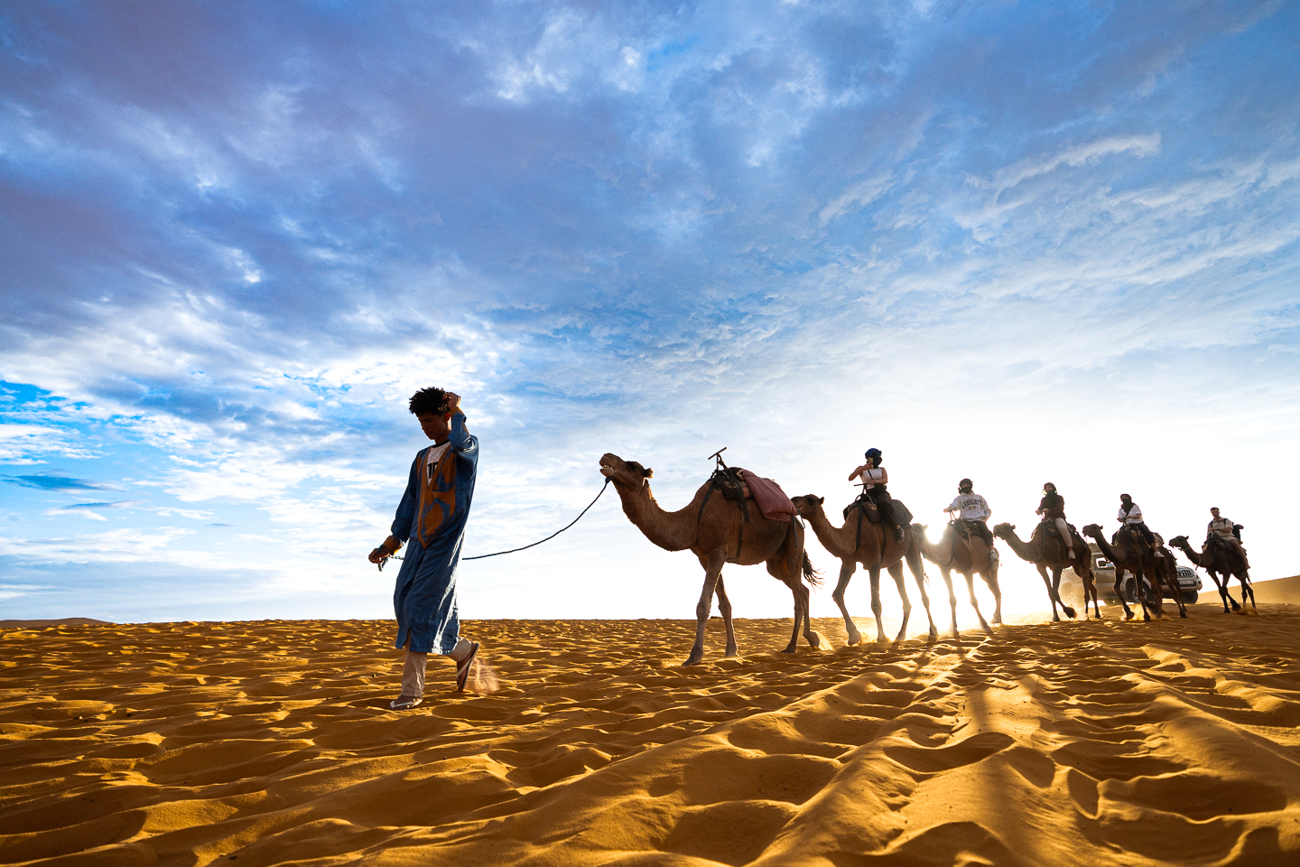 A guide in a blue robe leading a camel caravan across golden sand in Merzouga, Morocco, with sunlight filtering through a cloudy sky.