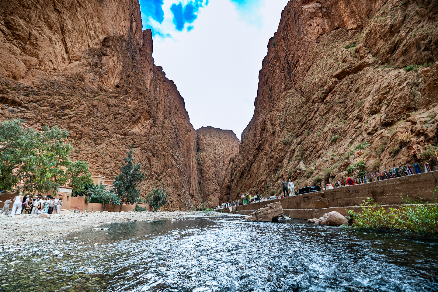 Shallow river flowing between towering rock walls in Todra Gorges, Morocco, with visitors along the rocky bank and roadside.